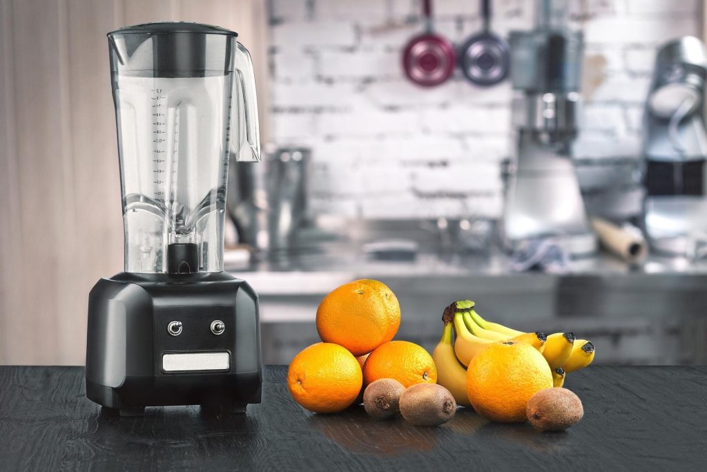 Blender med glaskanna bredvid apelsiner, bananer och kiwi på en köksbänk