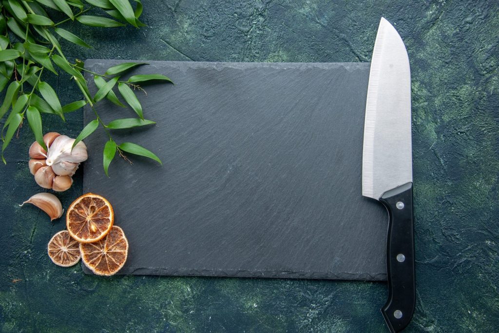 Kockkniv bredvid en mörk skärbräda av sten med vitlök och torkad citrus