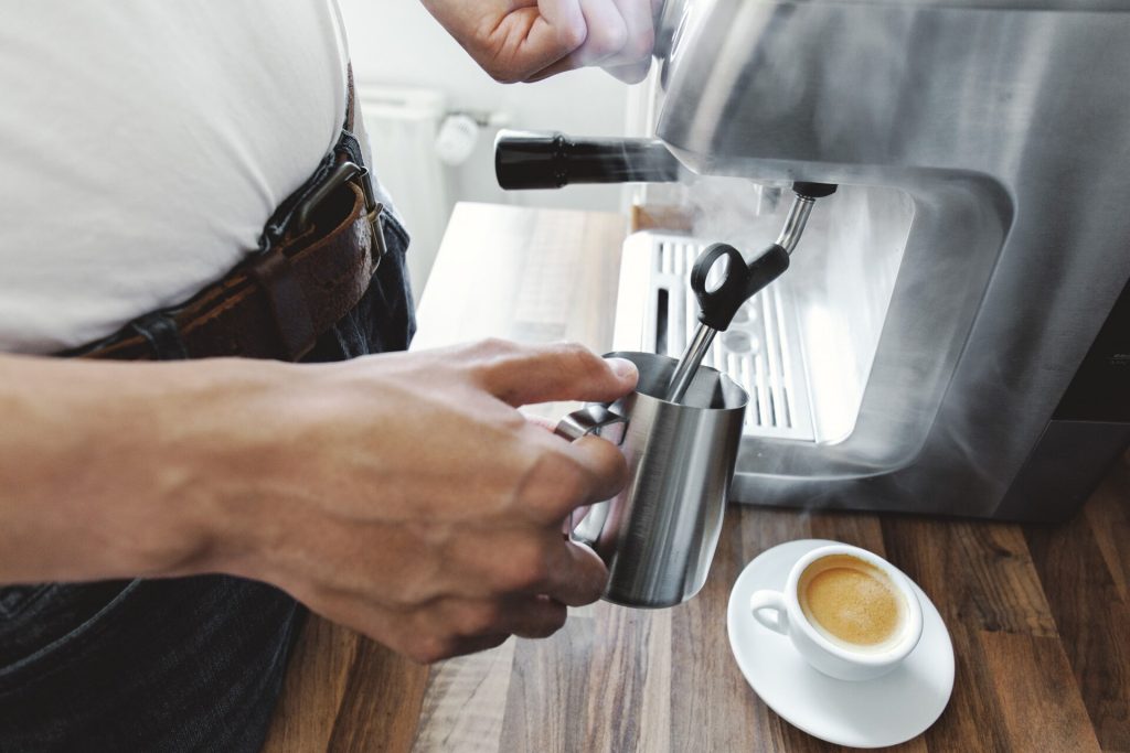 Person skummar mjölk i en kanna vid en kaffemaskin bredvid en espressokopp