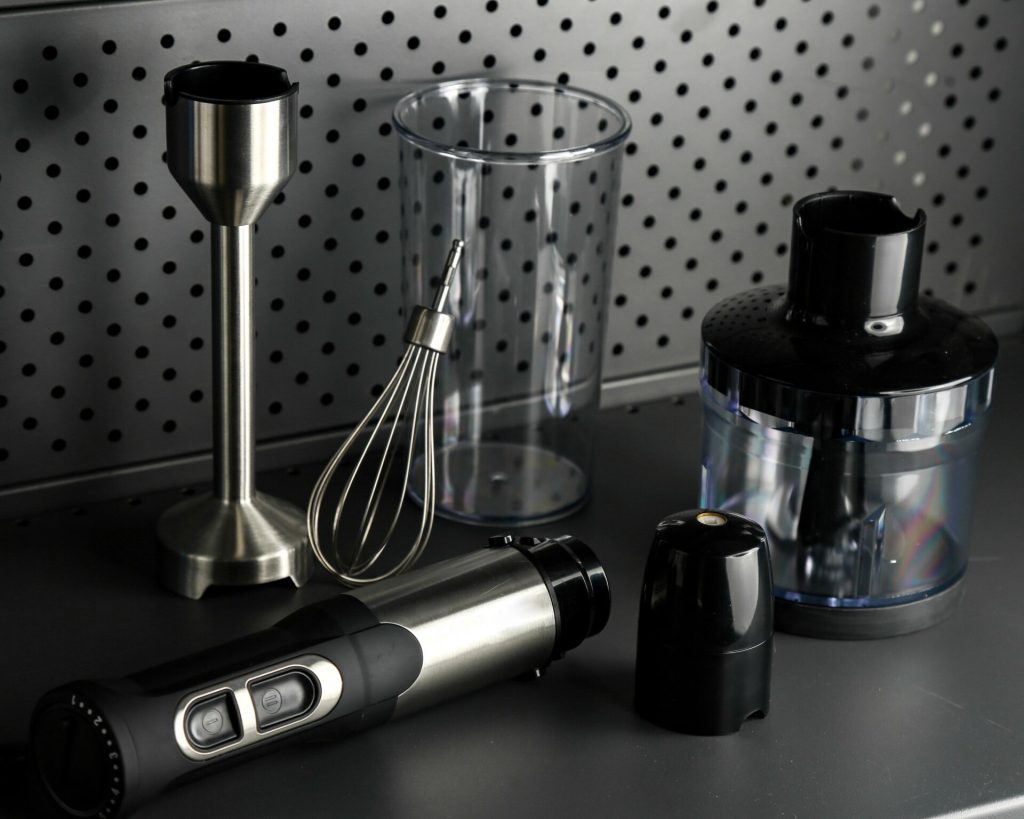 Stavmixer med visp, mixerbägare och hackare på en mörk köksbänk